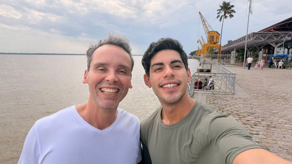 Dois homens sorrindo juntos em frente a um corpo d'água, com um cais e palmeiras ao fundo.